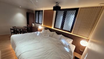 A hotel room with a large bed, a desk, and a chair. There are windows with wooden frames and a ceiling fan.