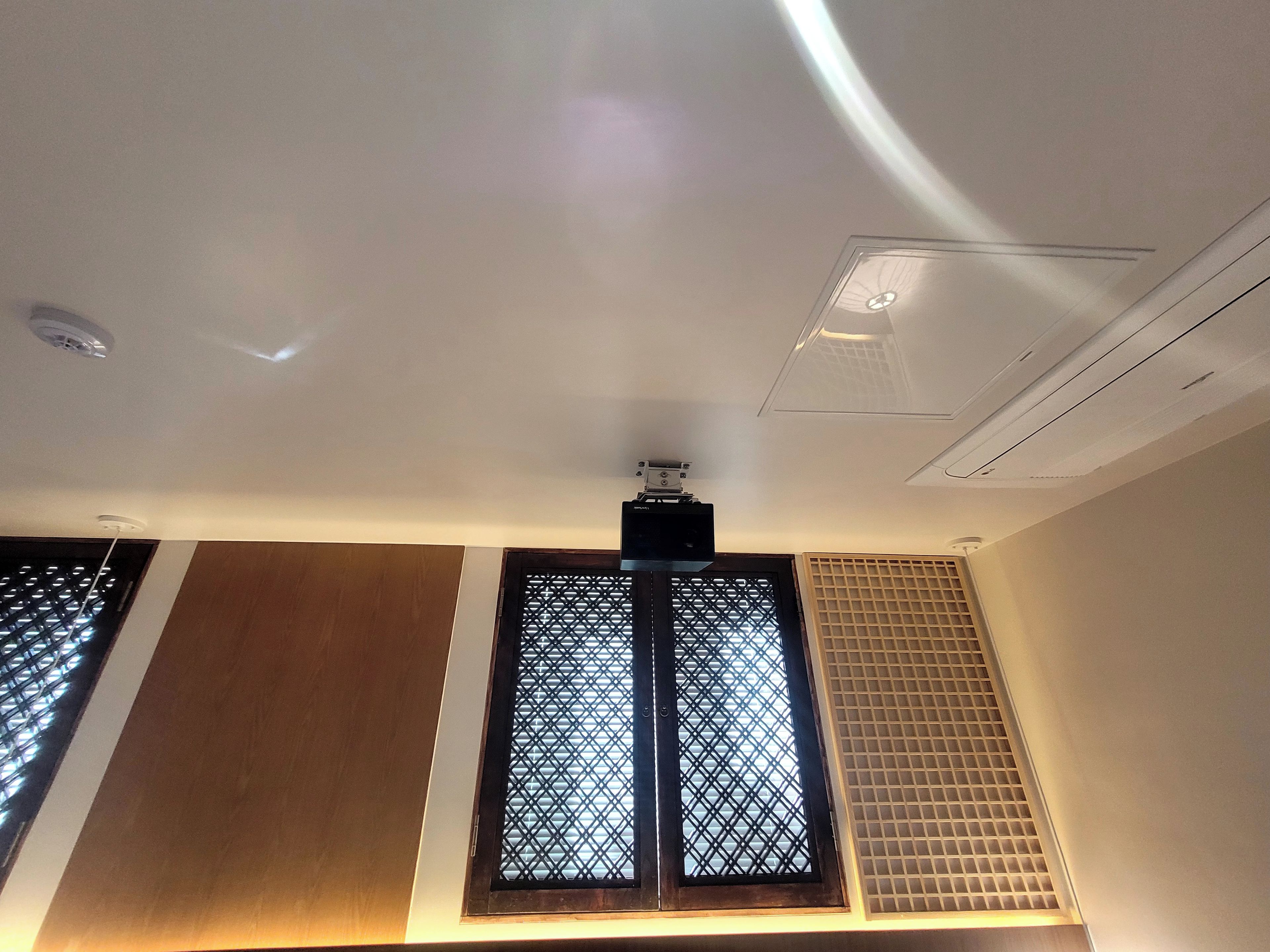 A room with a ceiling-mounted air conditioning unit, a window with a grid pattern, and a wall with a wooden panel.