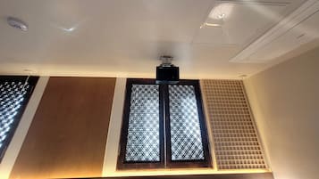 A room with a ceiling-mounted air conditioning unit, a window with a grid pattern, and a wall with a wooden panel.