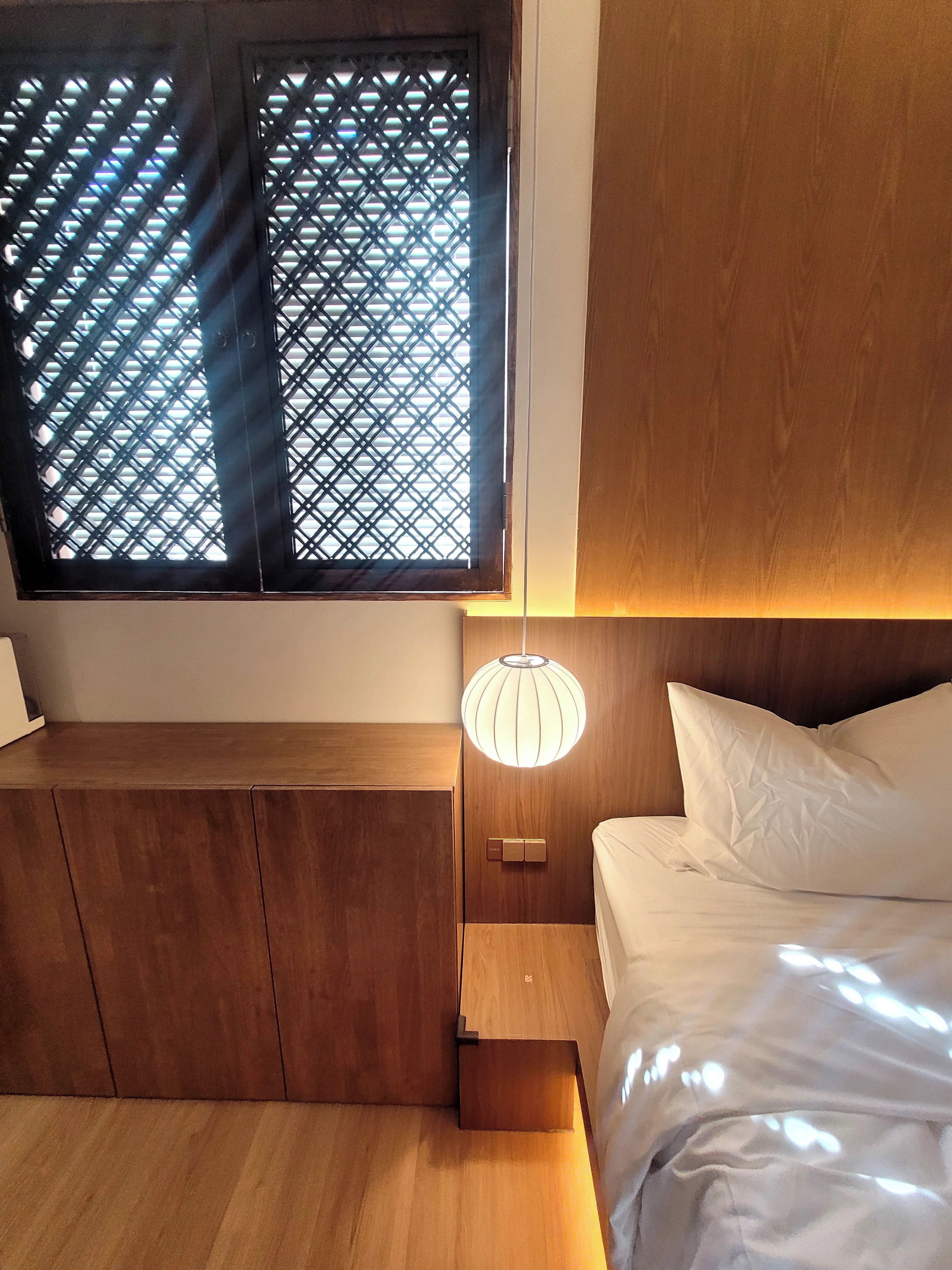 Un dormitorio con una cama, una mesita de noche, una lámpara de pared y una ventana con diseño de celosía.
