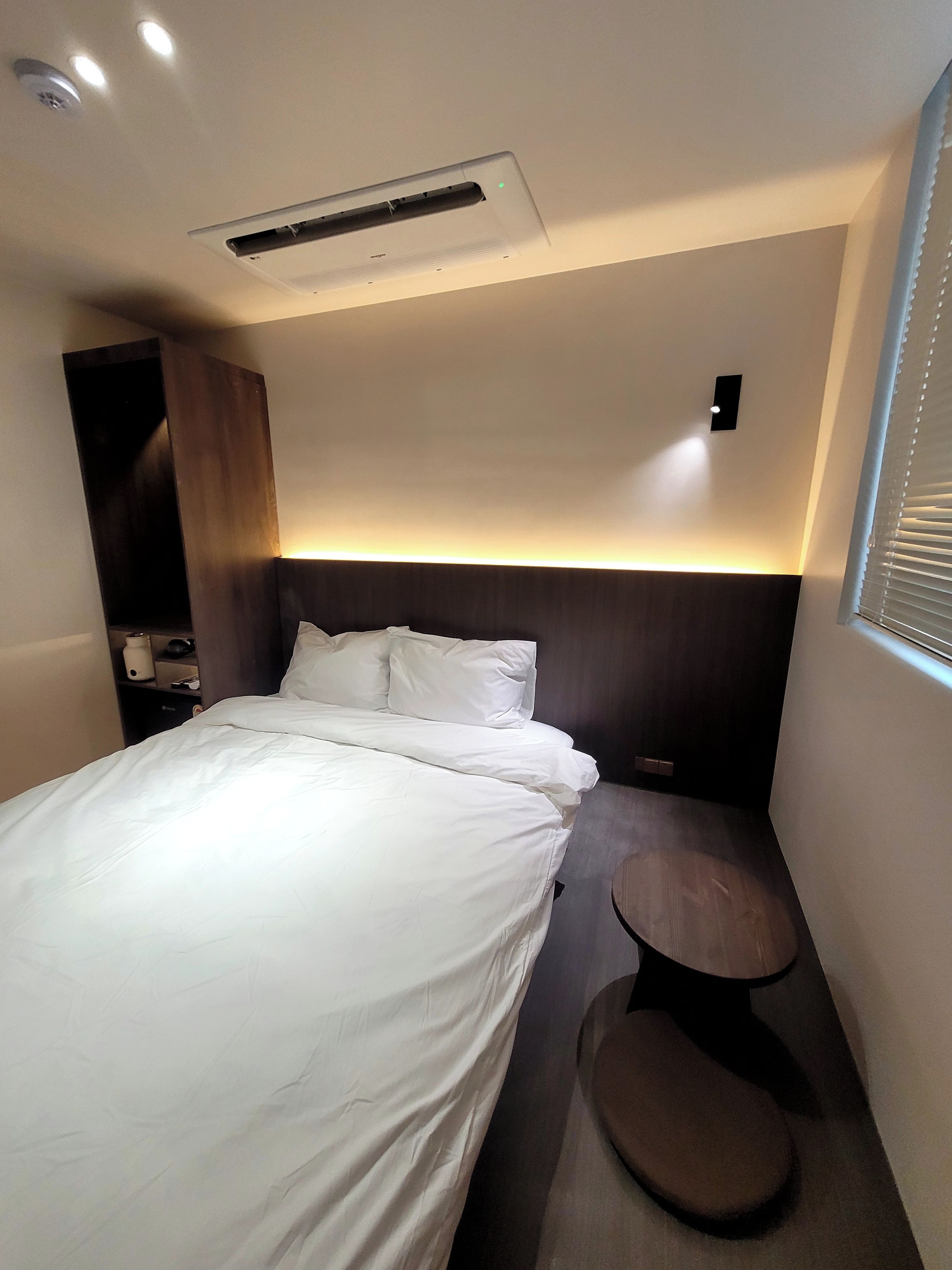 A hotel room with a bed, bedside table, and a wall-mounted air conditioning unit.