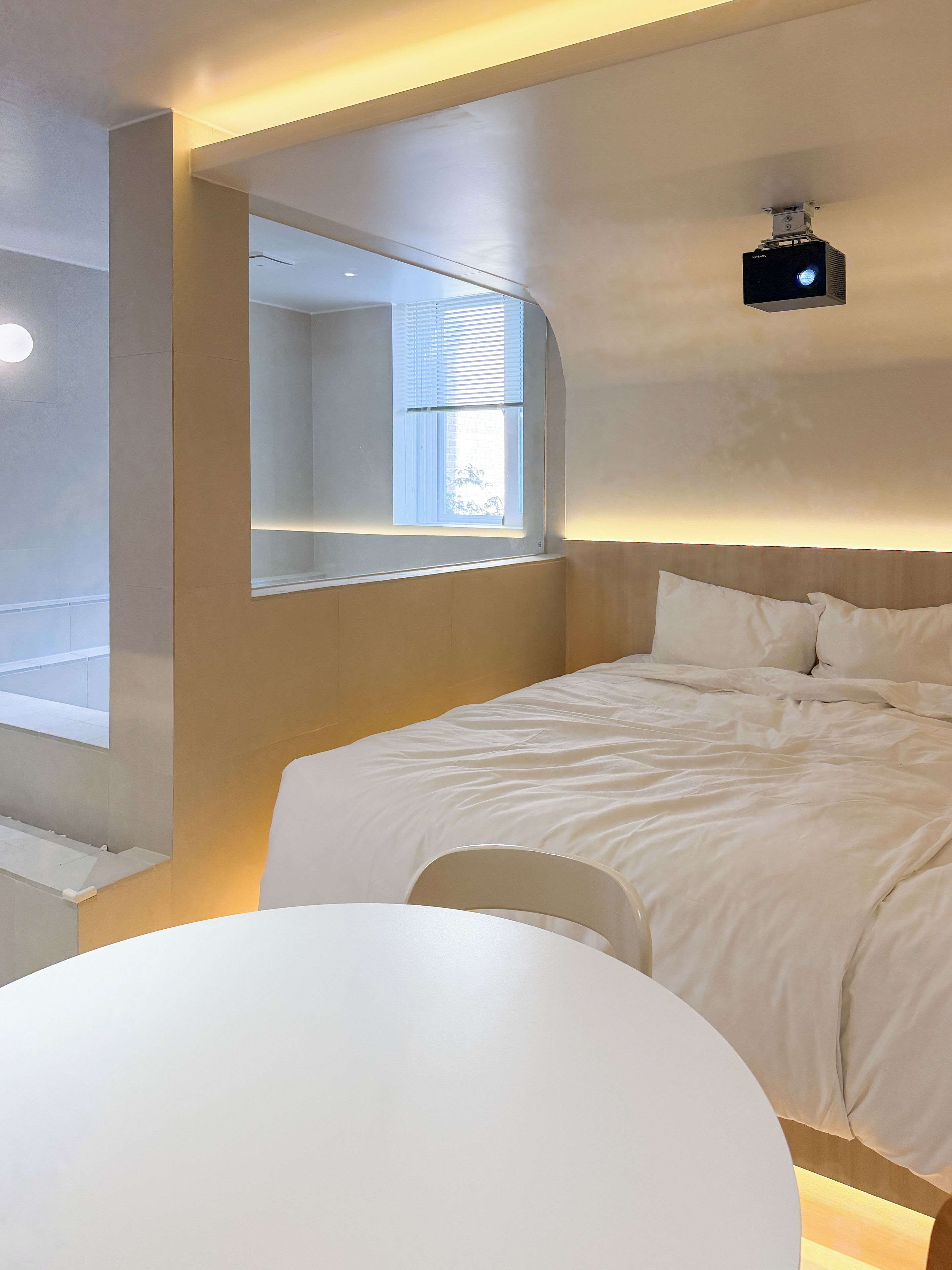 A bedroom with a bed, a bedside table, a chair, a window, and a ceiling-mounted projector.