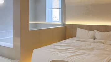 A bedroom with a bed, a bedside table, a chair, a window, and a ceiling-mounted projector.