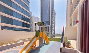 A playground with yellow and green equipment, surrounded by modern buildings with reflective glass windows. - Vogue | VYB at Business Bay close to Downtown (Dubai)