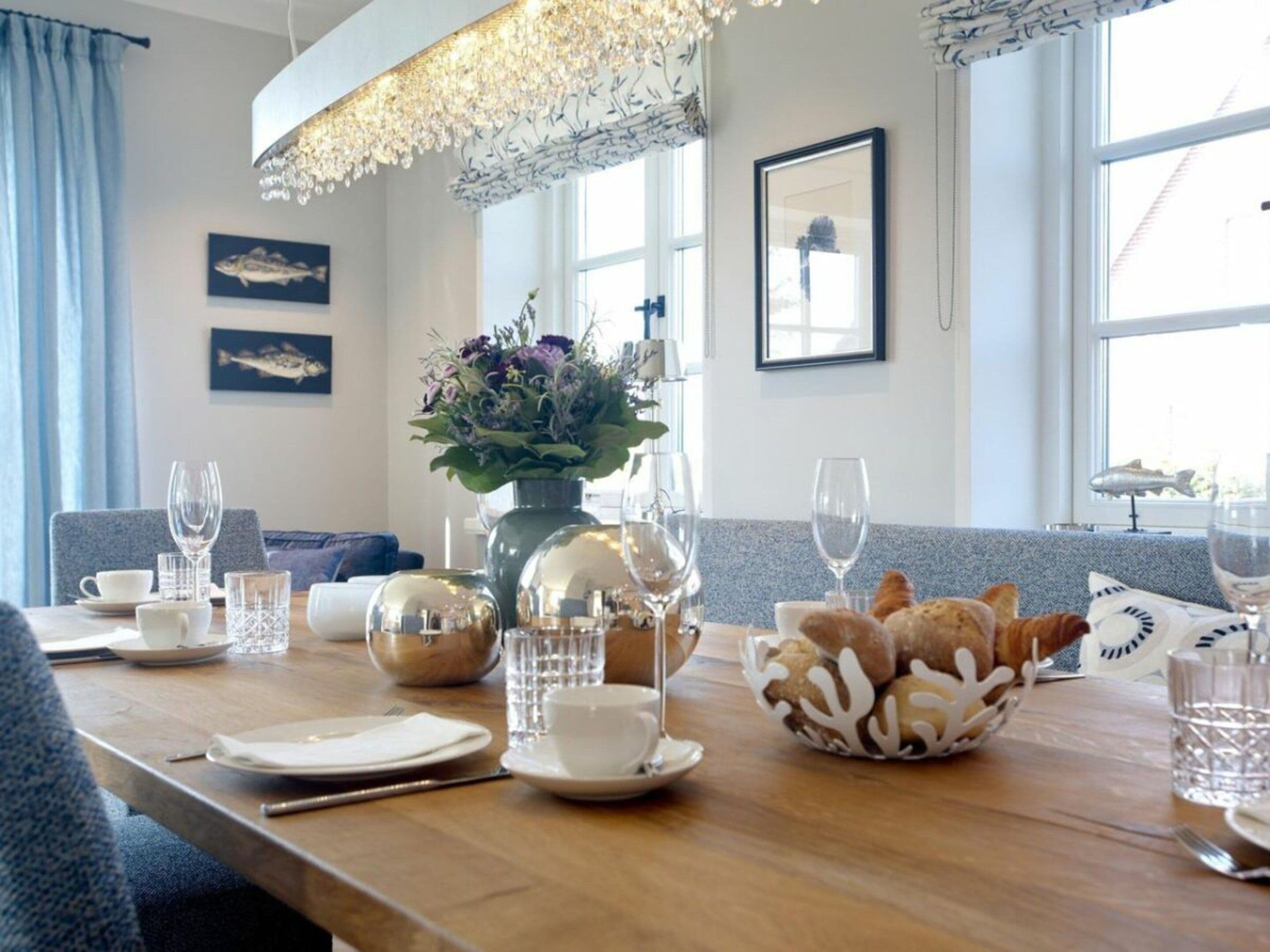 A dining area with a wooden table set for two, featuring clear glassware, a vase with flowers, and a bowl of bread.