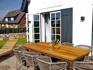 Une terrasse extérieure avec une table en bois et des chaises en osier, un mur de pierre et une maison à l’extérieur blanche et à la porte sombre.
