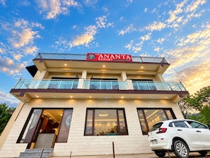Un hotel con un letrero que dice 'The Ananta Resort'. Tiene varios pisos, balcones con barandillas de vidrio y un coche blanco estacionado adelante.