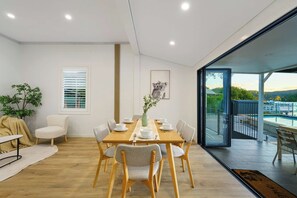 A dining area with a wooden table and chairs, a vase with greenery, and a view of a pool and landscape through a glass door. - Waterfront Escape with Jetty and Pool (Woy Woy)