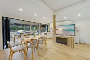 A modern living room with a dining area, a flat-screen TV, and a view of the outdoors. - Waterfront Escape with Jetty and Pool (Woy Woy)