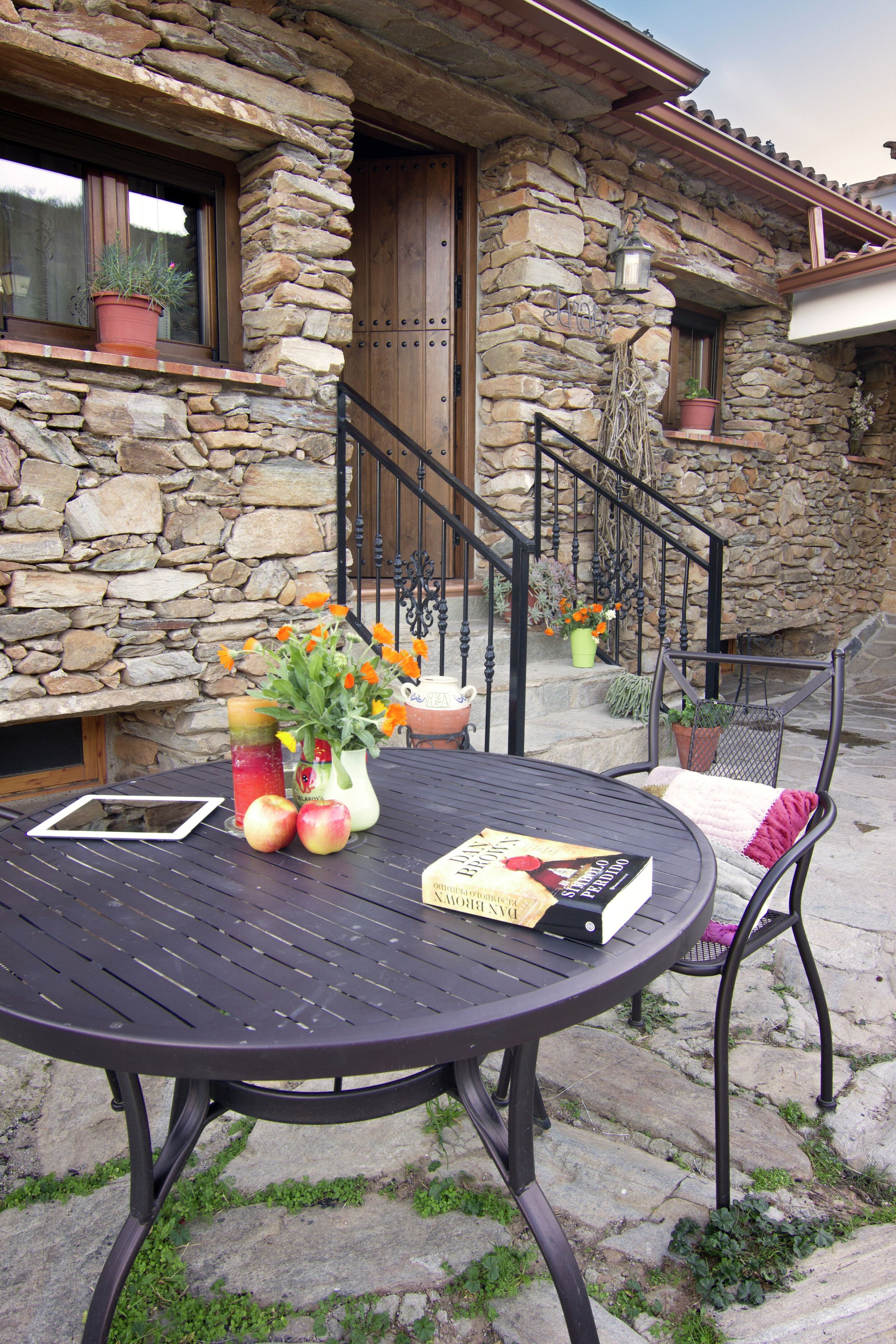Una casa de piedra con puerta de madera, una escalera y una mesa redonda con un libro y fruta.