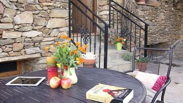 Una casa de piedra con puerta de madera, una escalera y una mesa redonda con un libro y fruta.