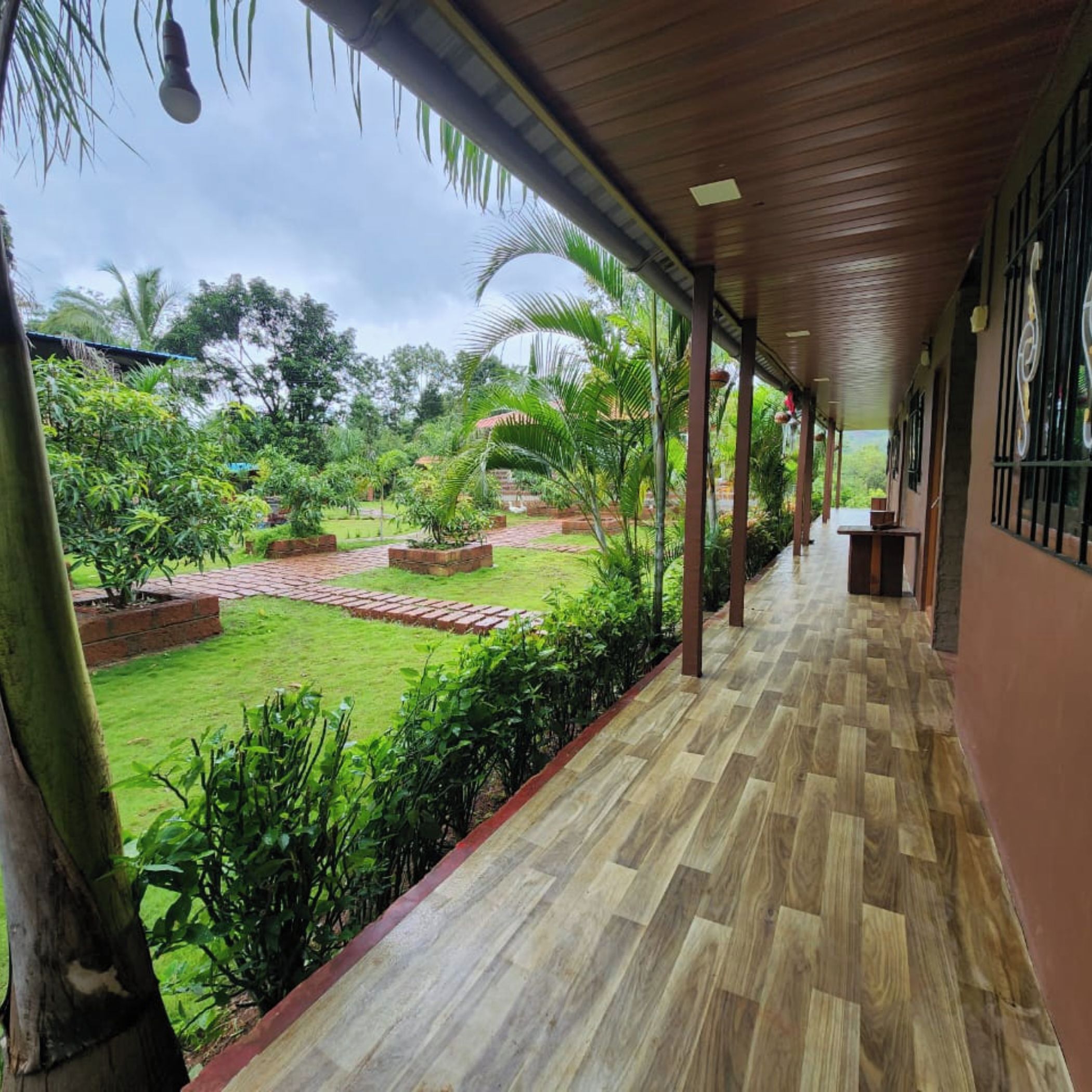 A covered walkway with a wooden floor, leading to a lush garden with various plants and a brick pathway.