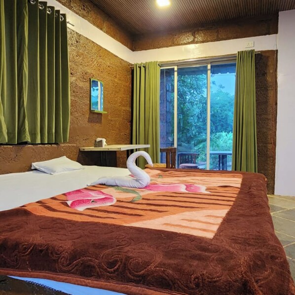A hotel room with a bed, a small table, a chair, a window with curtains, and a view of greenery outside.