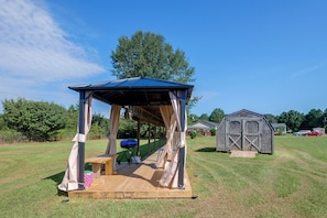 A covered outdoor seating area with a bench and a grill, a wooden deck, and a small shed in a grassy field.