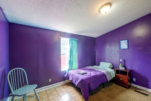 Ein Schlafzimmer mit violetten Wänden, einem Bett mit violetter Bettdecke, einem Sessel, einem Nachttisch mit Lampe und einem Fenster mit Vorhängen.
