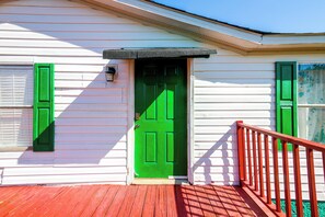 A white building with a green door and matching shutters, a red deck, and a black awning.