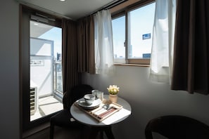 A small dining area with a table set for two, a chair, a vase with flowers, and a view of the city through a window. - JY Suites Namba (Nipponbashihigashi, Naniwa Ward)