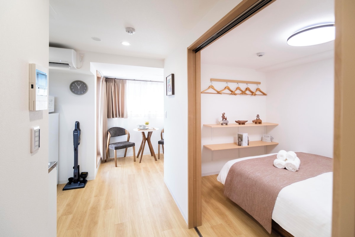 A hotel room with a bed, a desk with a chair, a vacuum cleaner, a clock, and a shelf with decorative items.