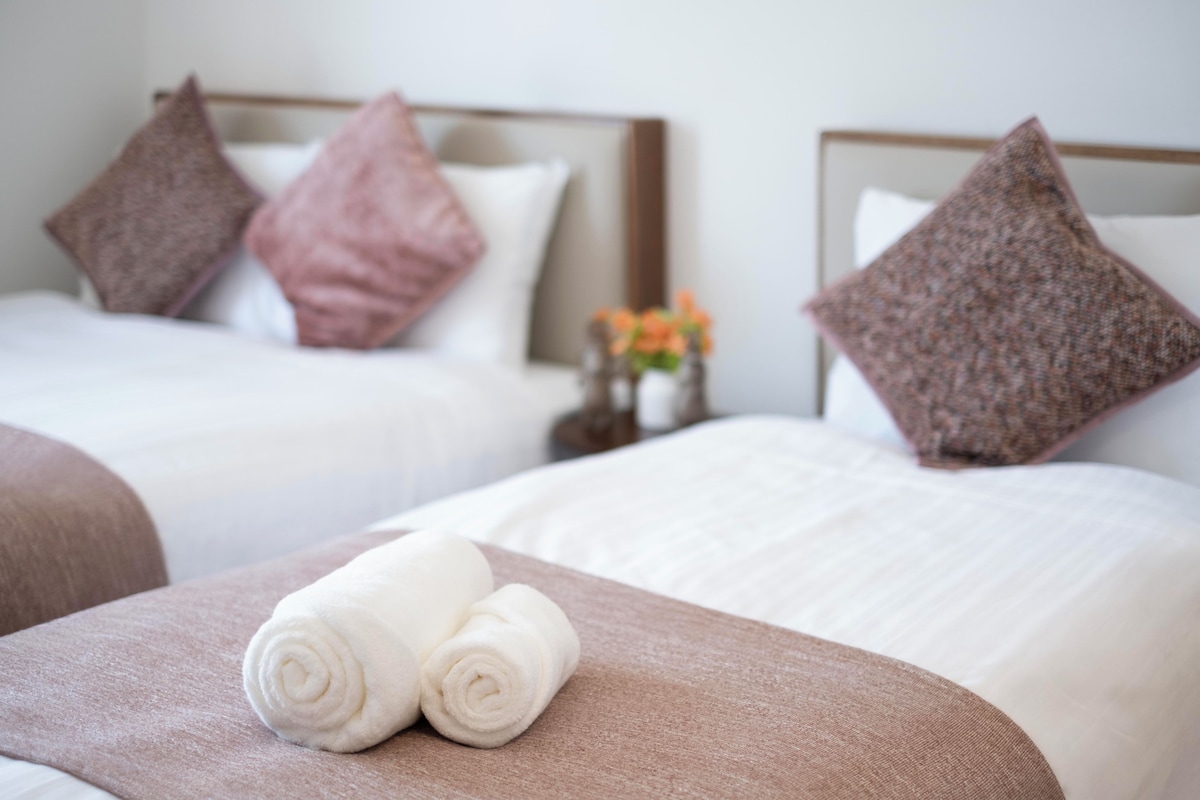 A hotel room with two beds, each with a headboard, pillows, and a rolled white towel on the bed.