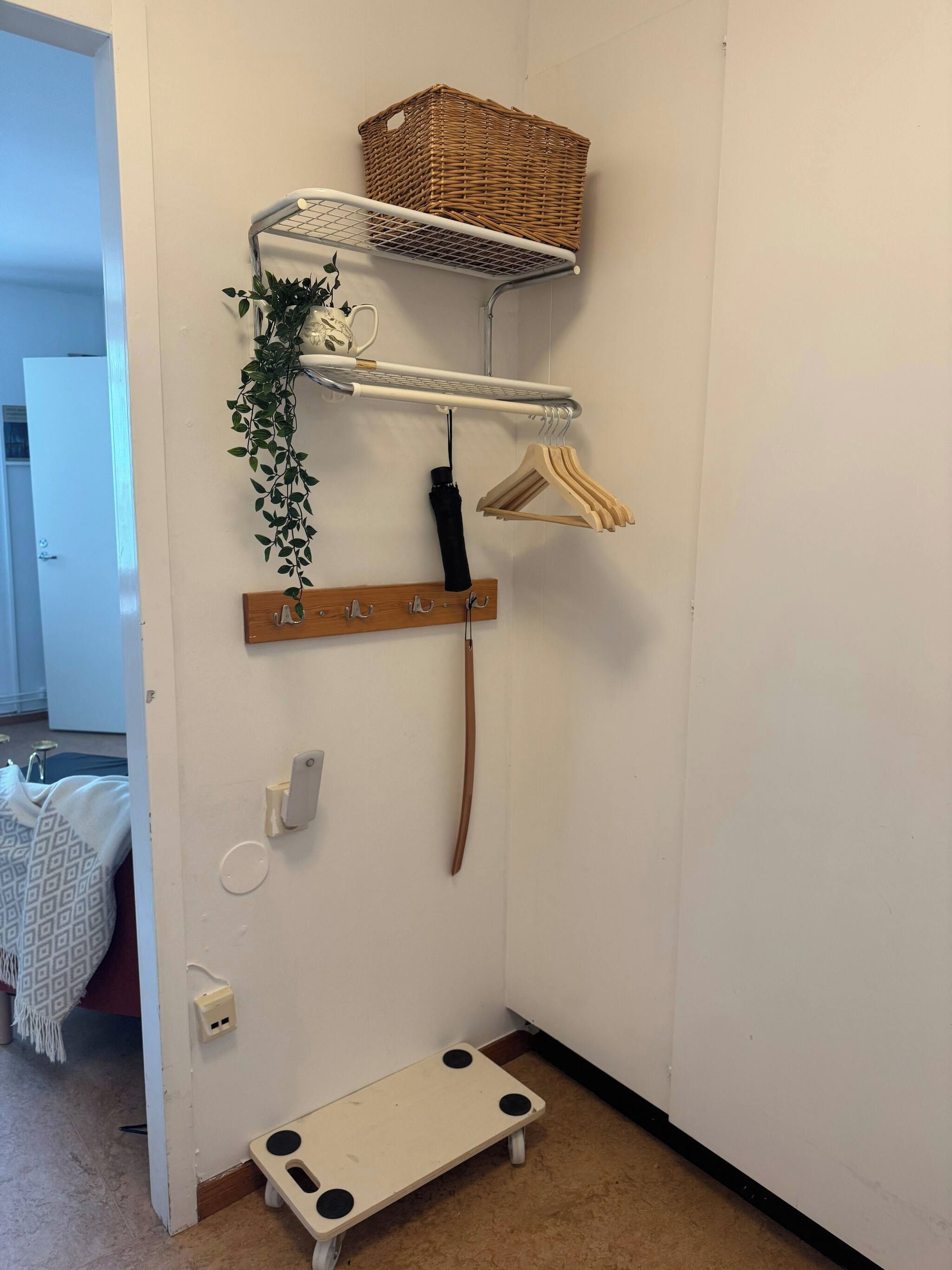 Canto de um quarto com parede branca, um pequeno banco de madeira, uma prateleira branca com um cesto de vime, um cabideiro com ganchos e uma planta pendente.