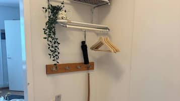 Canto de um quarto com parede branca, um pequeno banco de madeira, uma prateleira branca com um cesto de vime, um cabideiro com ganchos e uma planta pendente.