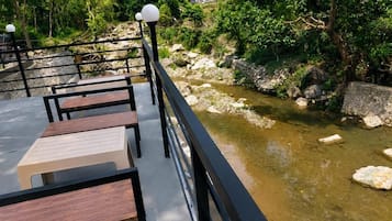 Eine Uferterrasse mit Holztischen und -stühlen, einem Geländer und Blick auf einen Fluss mit Felsen und Grünflächen.
