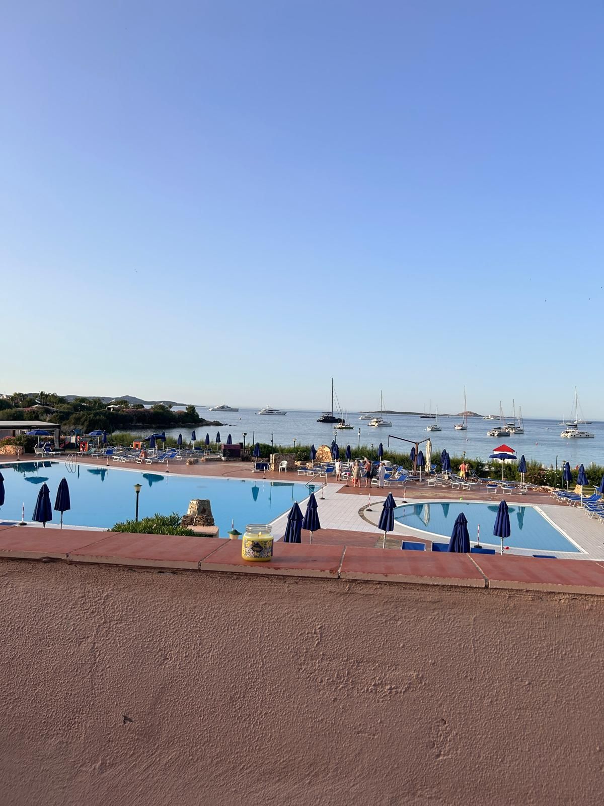 Een zwembad met ligbedden en parasols, omringd door een strand en boten op de achtergrond.
