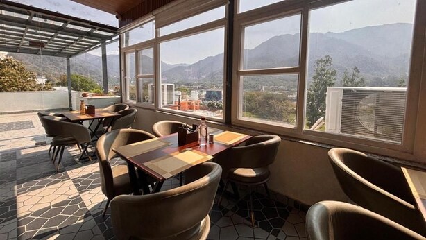 An indoor seating area with tables and chairs, large windows with a mountain view, and a tiled floor. - Hotel Shivay Palace (Rishikesh)