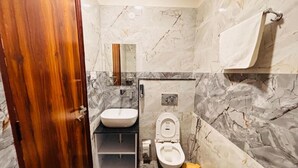 A bathroom with a marble wall, a wooden door, a sink, a toilet, and a trash bin. - Hotel Shivay Palace (Rishikesh)
