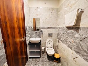 A bathroom with a marble wall, a wooden door, a sink, a toilet, and a trash bin. - Hotel Shivay Palace (Rishikesh)