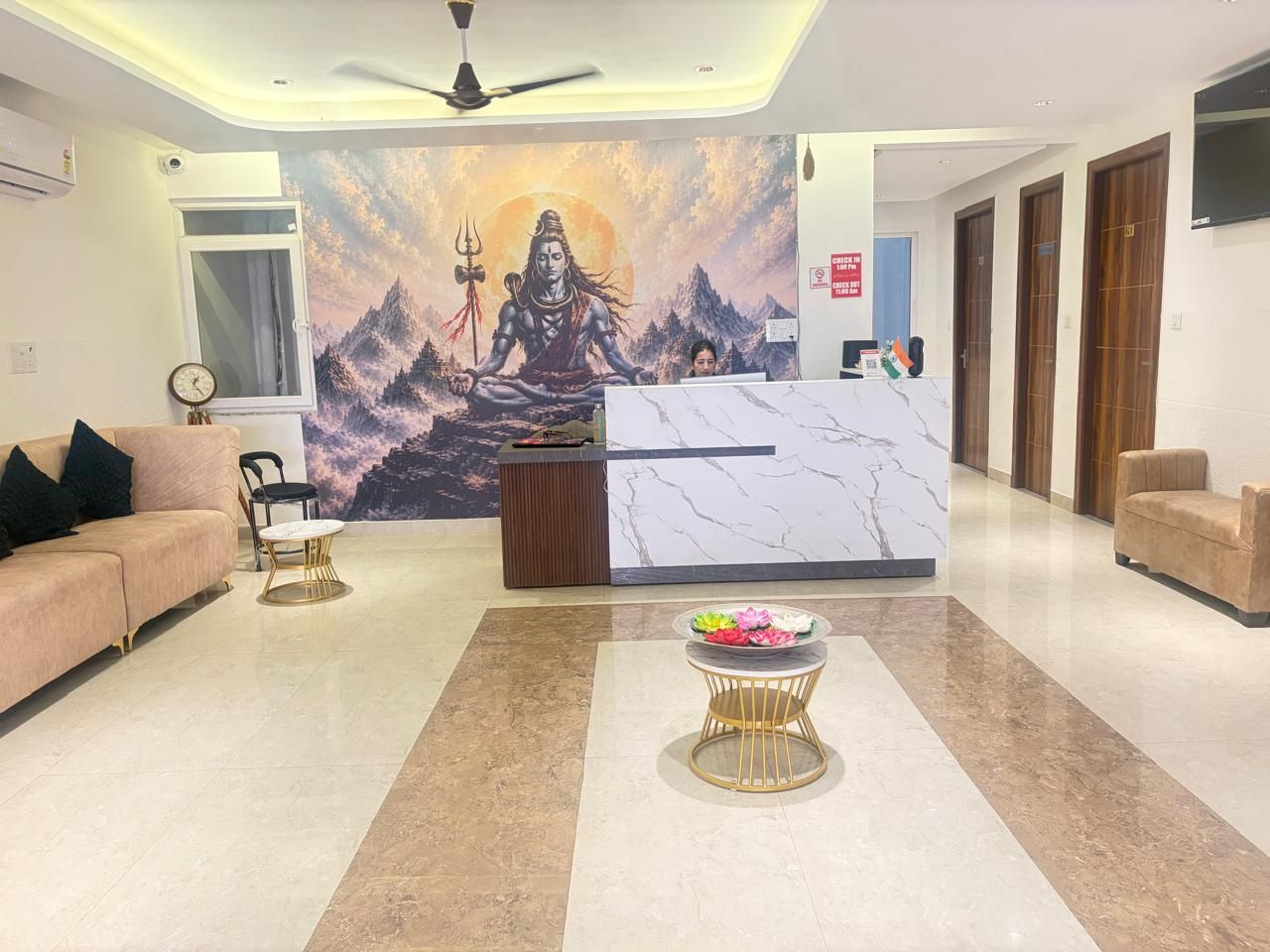 A hotel reception area with a marble reception desk, a sofa, a small round table with flowers, and a mural of a deity on the wall.