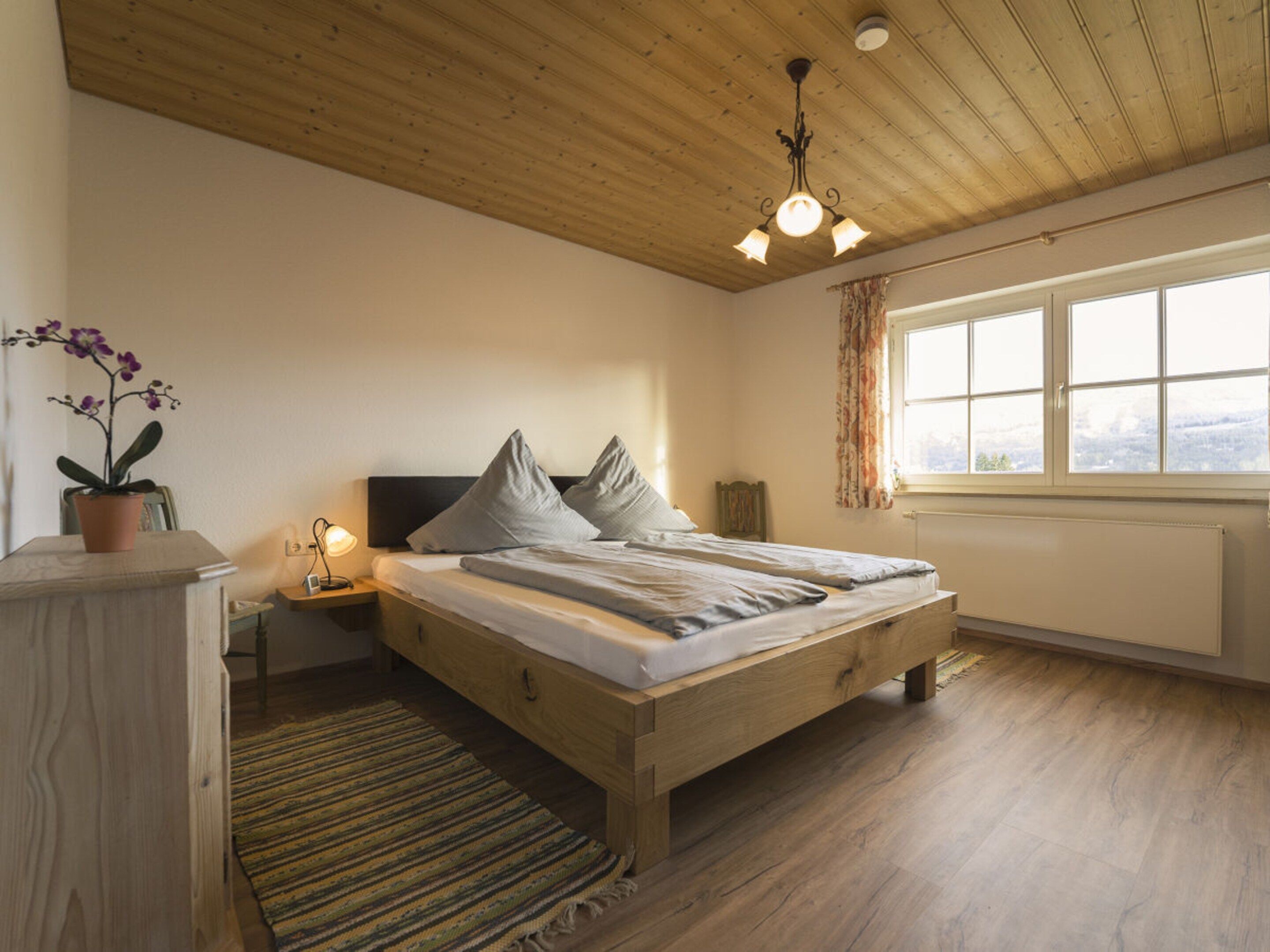 Ein Schlafzimmer mit einem hölzernen Bett, einem Nachttisch, einer Lampe, einem Fenster mit Vorhängen und einer Orchidee im Blumentopf.