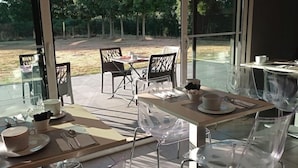 Un área de comedor al aire libre con mesas y sillas, vista a un campo cubierto de césped y un edificio con grandes ventanales.