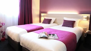 Habitación de hotel con dos camas, cada una con colcha blanca y morada, una revista sobre la primera cama y una mesita de noche con un teléfono.