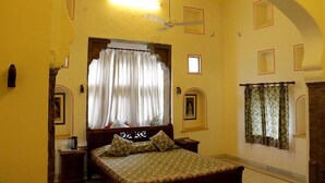 Una habitación con una cama, ventilador de techo, ventana con cortinas y una pared con cuadros enmarcados.