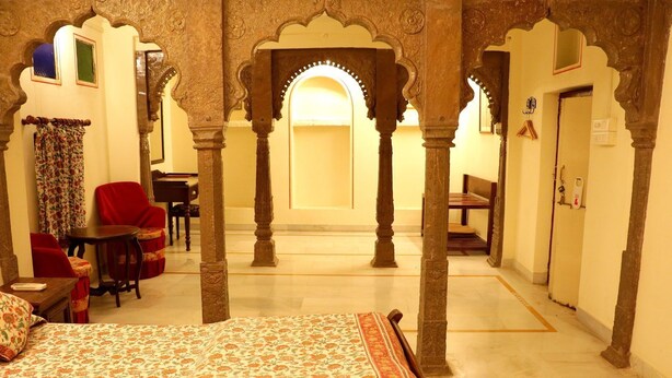 Una habitación con cama, una mesa, sillas y un escritorio pequeño. Se observan arcos y columnas en la arquitectura.