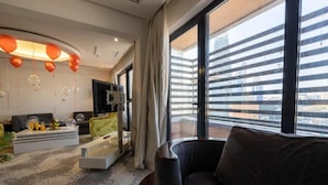 A modern living room with a sofa, a coffee table, and a TV. There is a large window with a view of buildings. - Mira Hotel (Riyadh)