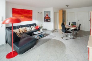 A modern living room with a black leather sofa, a red floor lamp, and a dining area with a table and chairs. - Holiday apartment with 1 bedroom (Norderney)