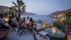 A group of people seated at tables outdoors with a view of the sea and hills. - Niriides Apartaments (Symi)