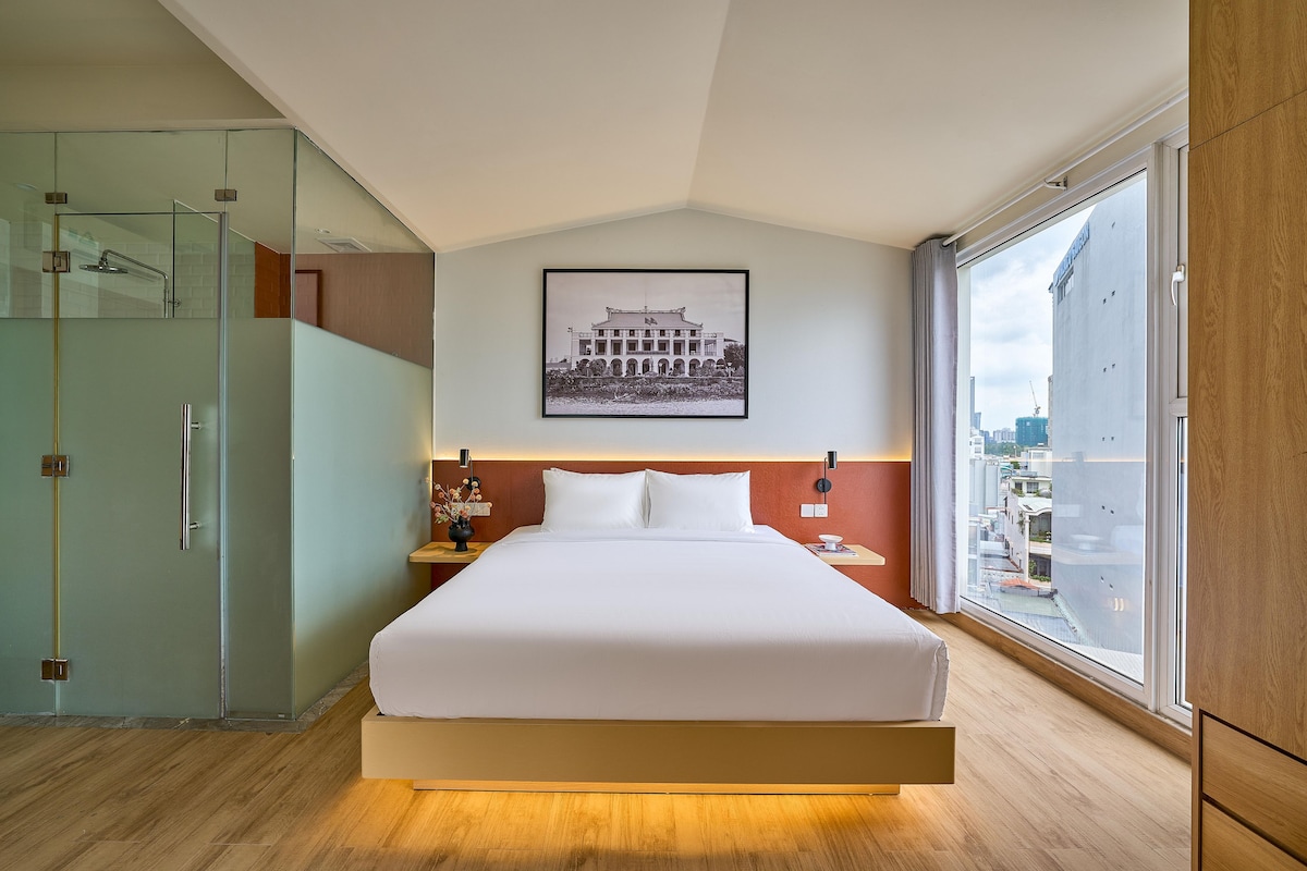 A modern hotel room with a large bed, a glass shower enclosure, a framed picture on the wall, and a city view through the window.