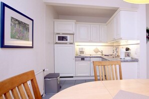 A compact kitchen with white cabinets, a microwave, and a sink. There is a framed picture on the wall and a chair at the table. - 6 at Gmelinstraße 23 (Wyk auf Föhr)