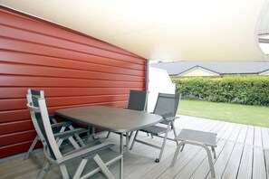 Um pátio externo com mesa e cadeiras, uma parede vermelha e vista para uma casa e um hedge.