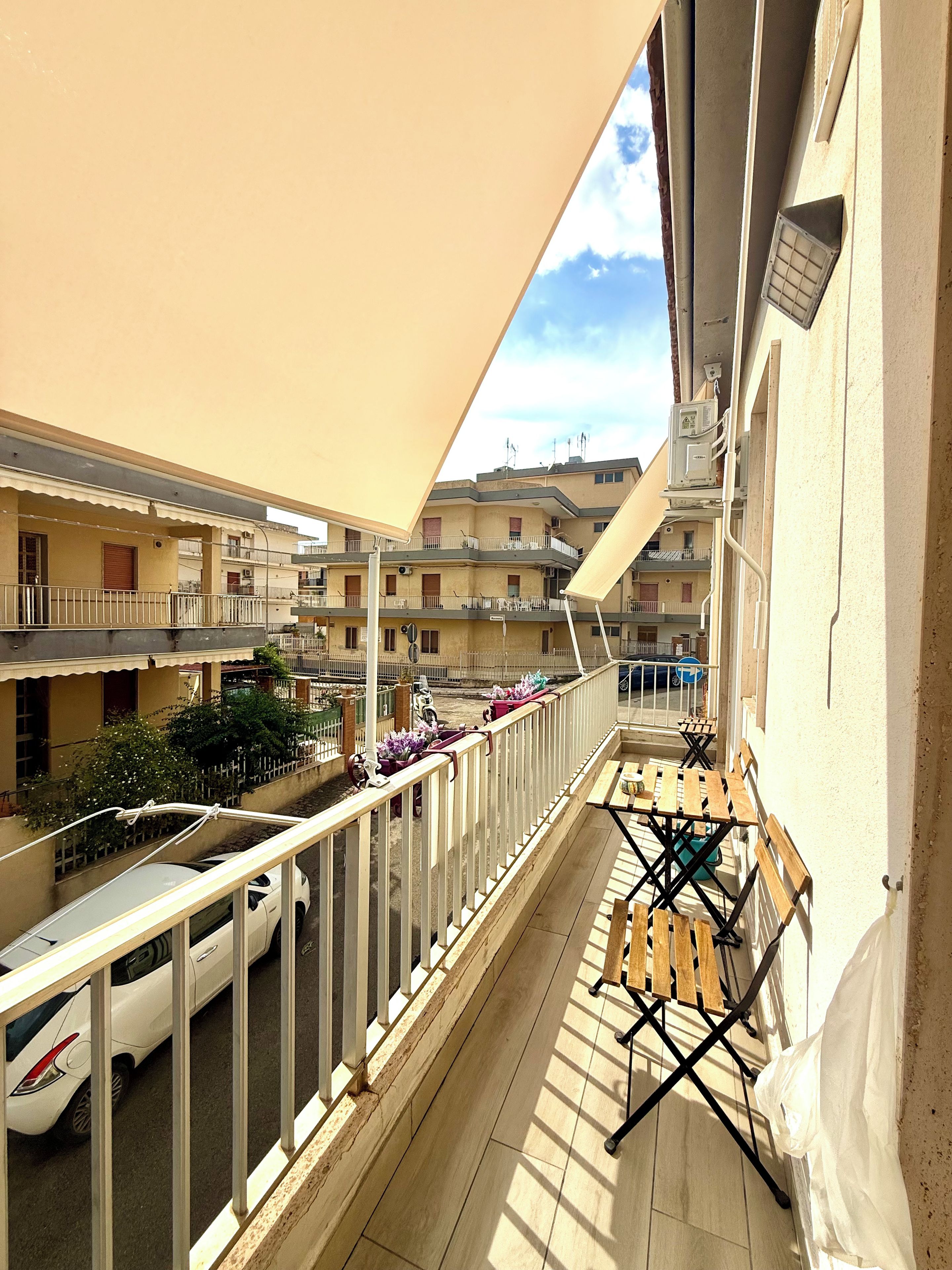 A balcony with a view of a multi-story building, a car parked on the street, and a wooden table with chairs.