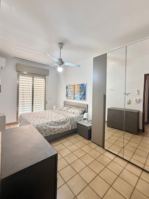A bedroom with a bed, bedside tables, a ceiling fan, a window with blinds, a painting on the wall, and a wardrobe. - Vacation rental sleeping 5 or 6 in the center of Marina di Ragusa. (Marina di Ragusa)