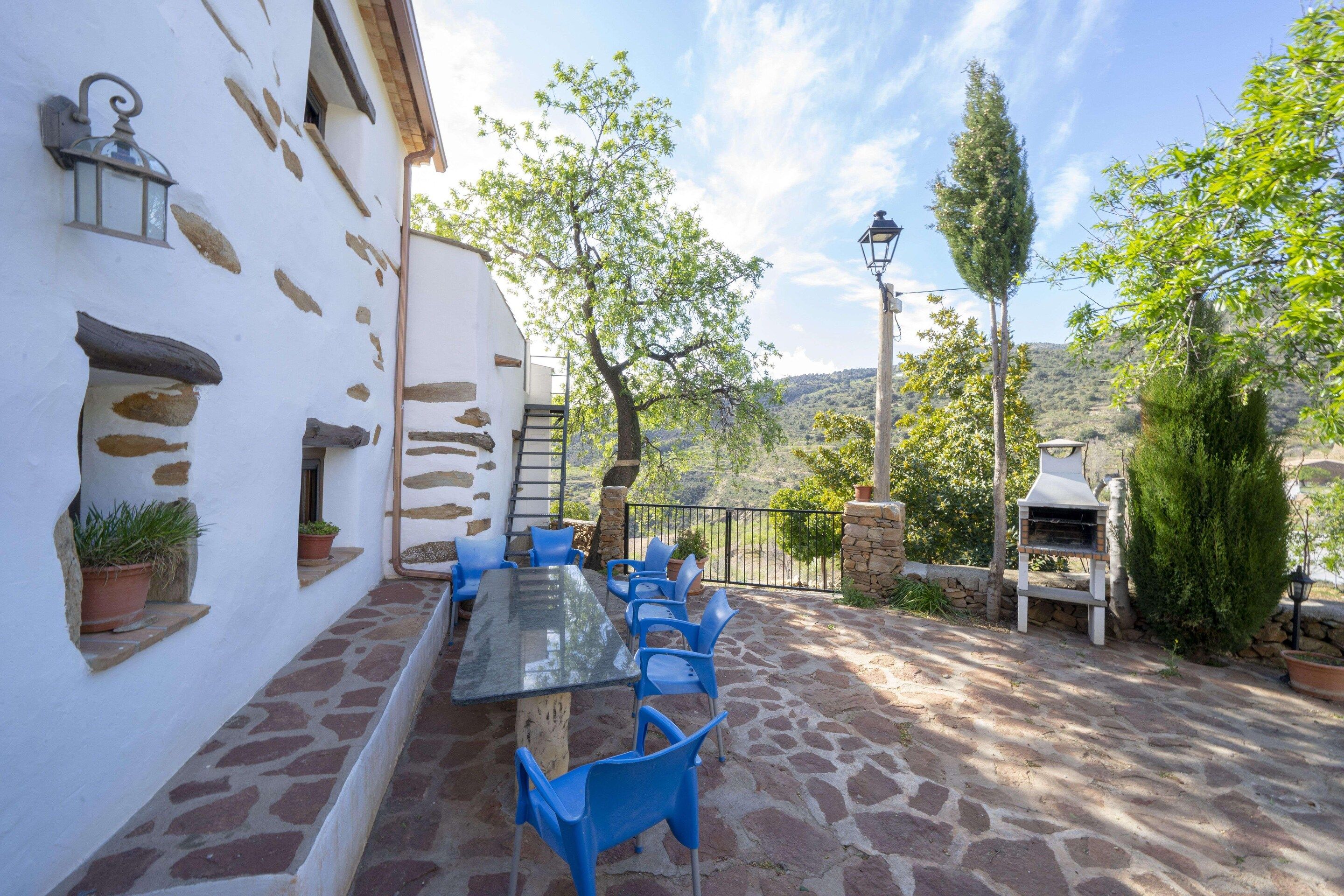 Un patio al aire libre con una mesa de vidrio y sillas azules, muros de piedra y vistas al campo.