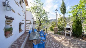 Un patio al aire libre con una mesa de vidrio y sillas azules, muros de piedra y vistas al campo.