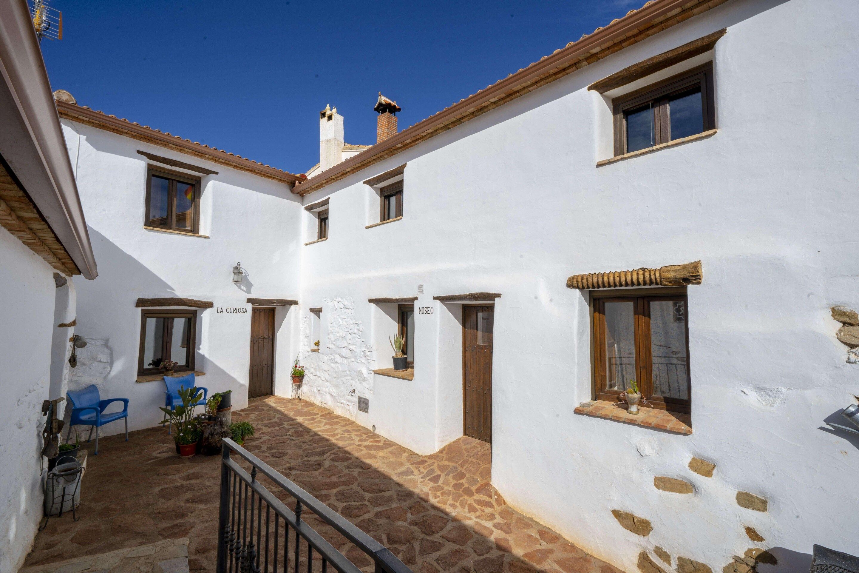 Un patio con paredes blancas, suelo de piedra y puertas de madera. Hay plantas en macetas y una silla azul.