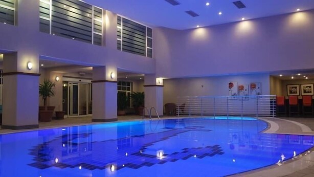 En indendørs swimmingpool med blå belysning på gulvet, omgivet af moderne arkitektur med store vinduer og et siddeområde med orange stole.