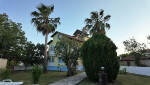 Una casa de dos pisos con un camino de entrada pavimentado, rodeada de palmeras y un césped cuidado.
