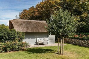Construção com telhado de palha, com um banco e uma pequena mesa, cercada por um jardim bem cuidado e uma parede de pedra.